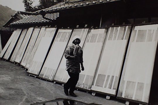 モノクロ写真:軒先に和紙を並べて乾かしているところ。ブラシを持った女性がこちらを見て微笑んでいる